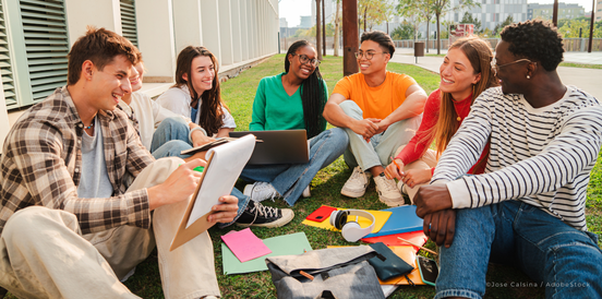 Eine bunte Gruppe Studenten sitzt zusammen auf dem grünen Rasen des Campus, während sie im Freien lernen, lesen und diskutieren, mit Notizbüchern, Laptops und Rucksäcken. - ©Jose Calsina/AdobeStock